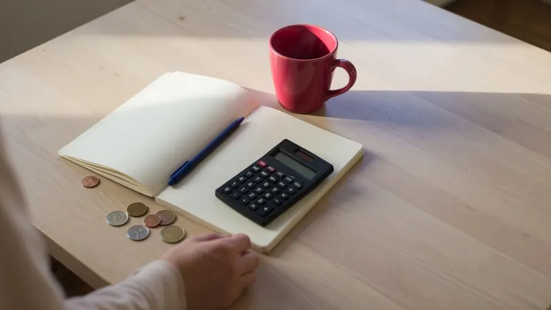 A kitchen table with a blank notebook, a calculator, a few coins and a raspberry coloured mug